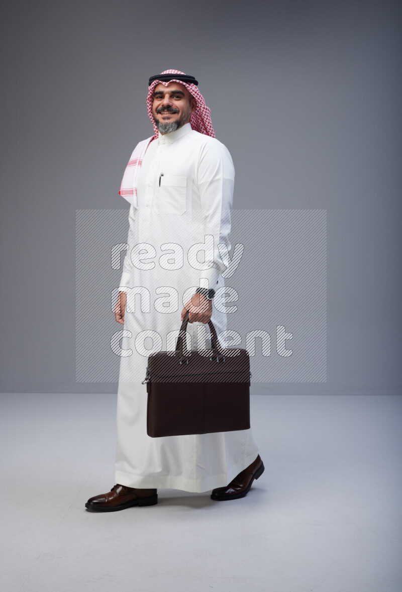 Saudi man Wearing Thob and red Shomag standing holding bag on Gray background