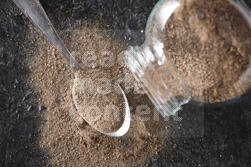A flipped glass spice jar full of black pepper powder with a metal spoon full of powder on textured black flooring
