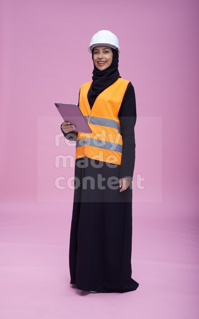 Saudi woman wearing Abaya with engineer vest and helmet standing working on tablet on pink background