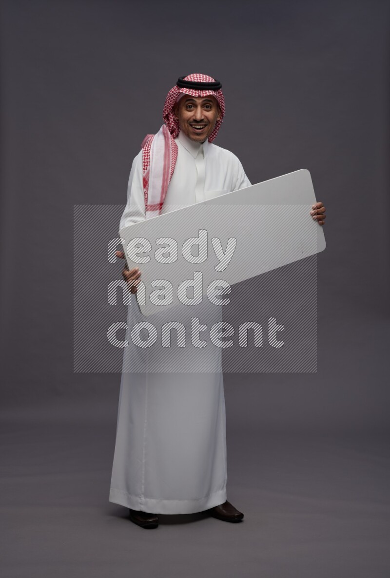 Saudi man wearing thob and shomag standing holding board on gray background