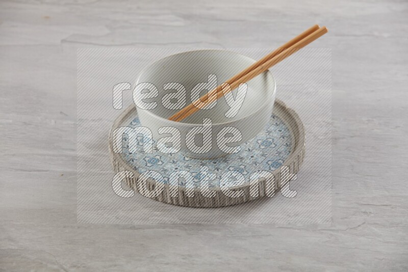 white pottery round bowl on top of multi color round ceramic plate and chopsticks, on grey textured countertop