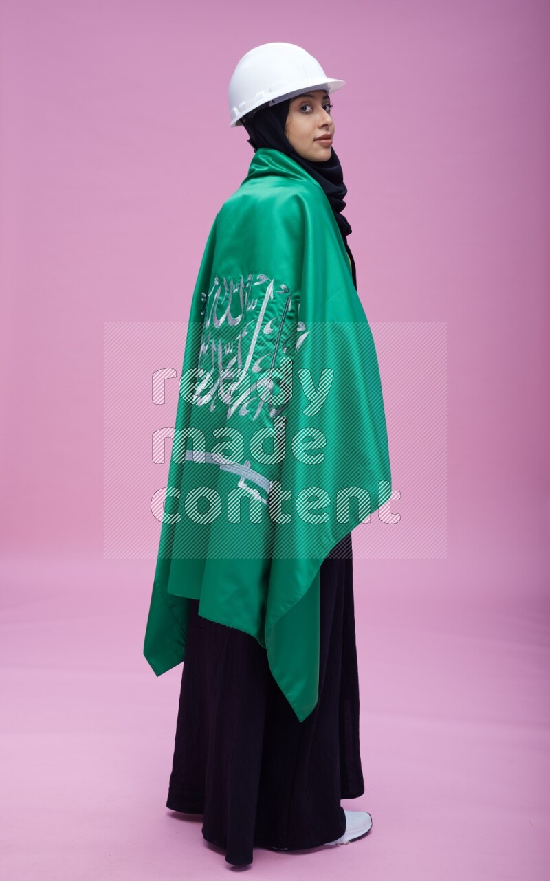 Saudi woman wearing Abaya with engineer vest and helmet standing holding Saudi flag on pink background