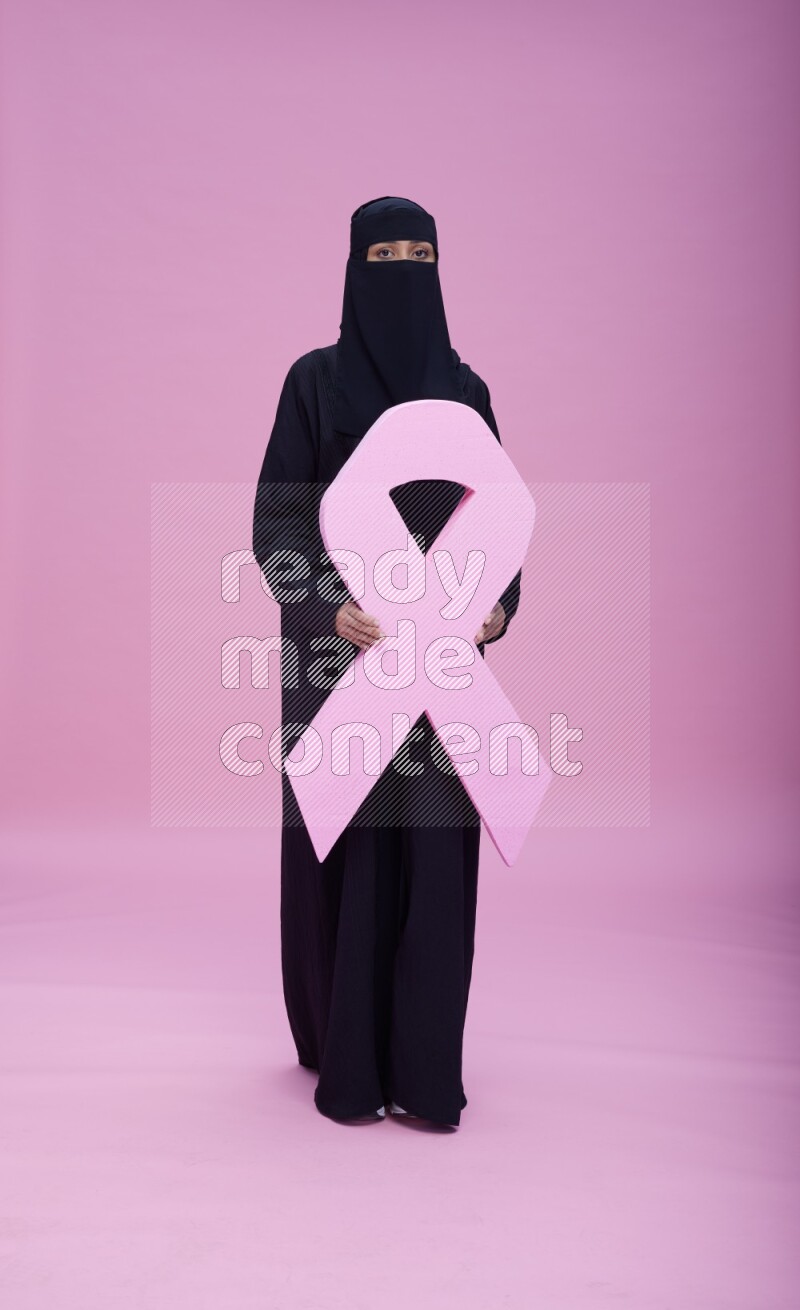 Saudi woman wearing abaya and niqab standing holding awareness ribbon on pink background