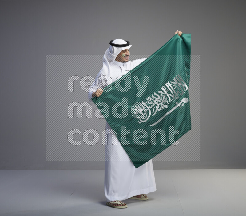 A Saudi man standing wearing thob and white shomag with face painting holding big Saudi flag on gray background