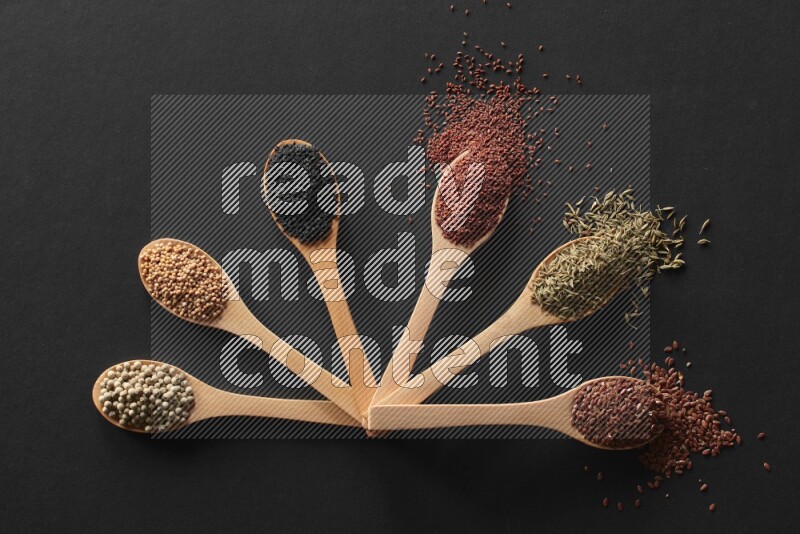 wooden spoons filled with white peppers, mustard seeds, black seeds, garden cress, cumin and flax on black flooring
