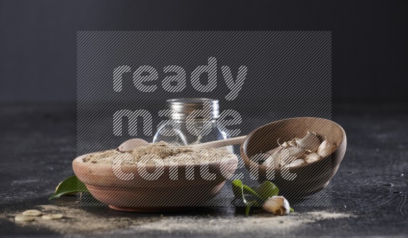 A wooden bowl with a spoon in it and glass spice jar all, full of garlic powder and a wooden bowl full of garlic cloves on a black flooring