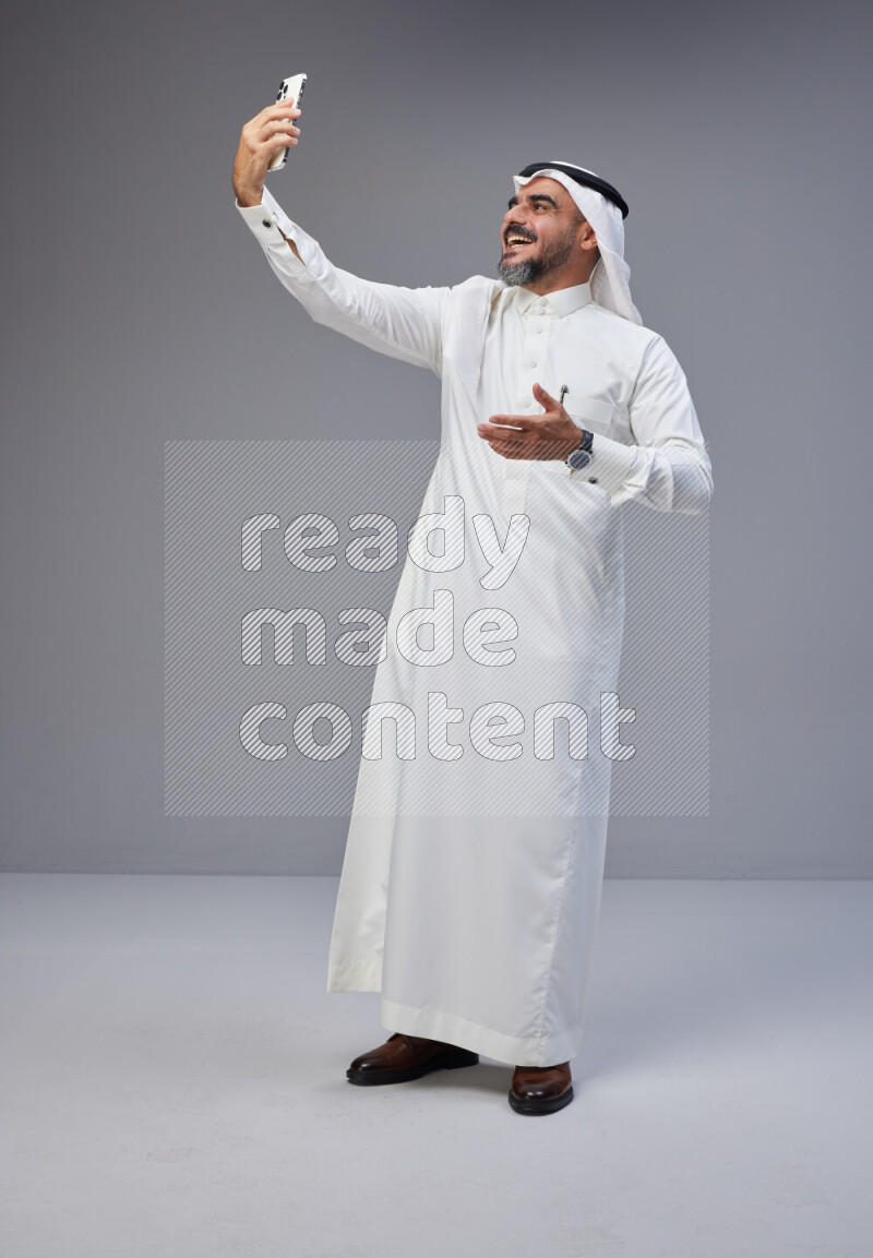 Saudi man Wearing Thob and white Shomag standing taking selfie on Gray background