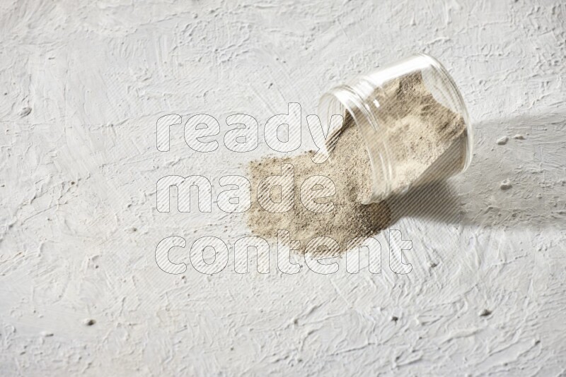 A flipped glass jar full of white pepper powder with spilled powder on textured white flooring