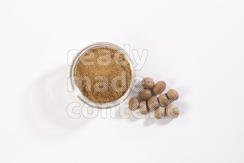 A glass bowl full nutmeg powder with the whole seeds beside it on a white flooring