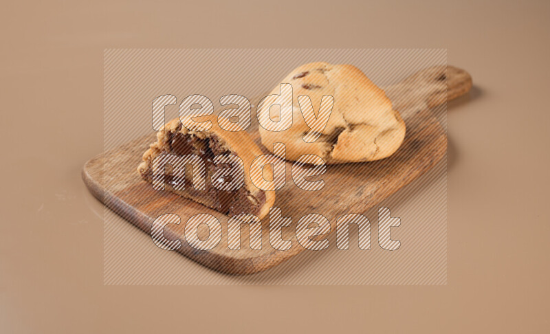 a chocolate chip cookie with another one cut in half on a wooden cutting board on a brown background