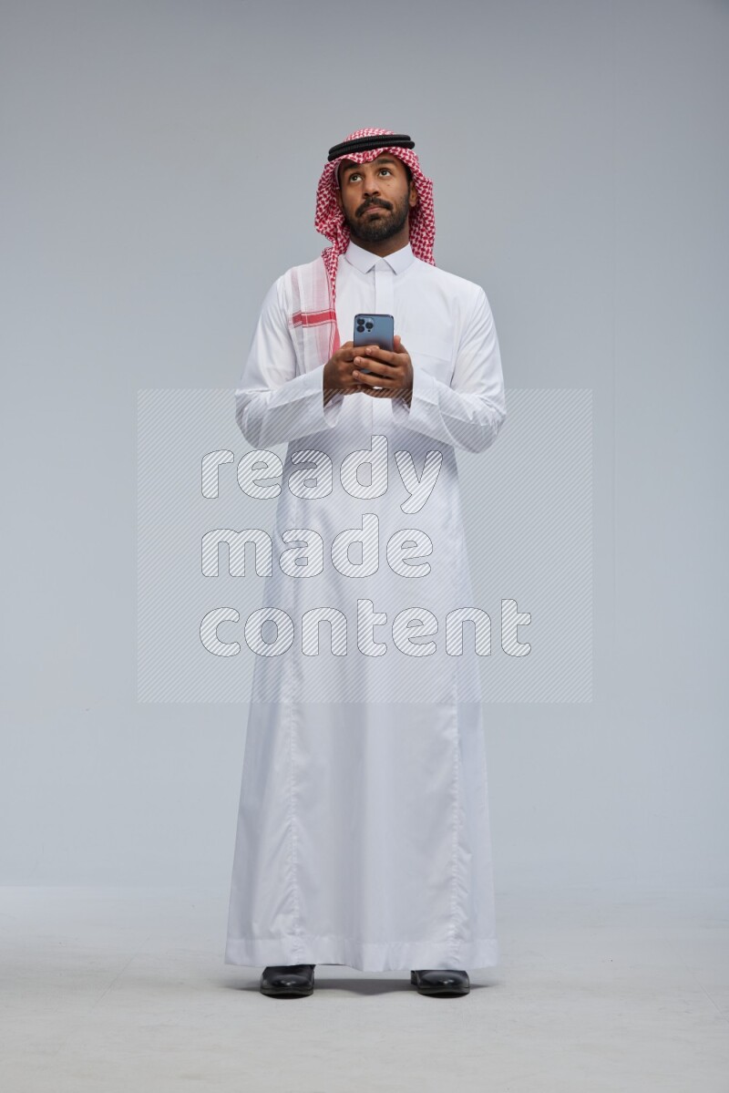 Saudi man Wearing Thob and shomag standing texting on phone on Gray background