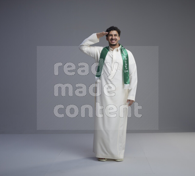 A Saudi man standing wearing thob and Saudi flag scarf on gray background