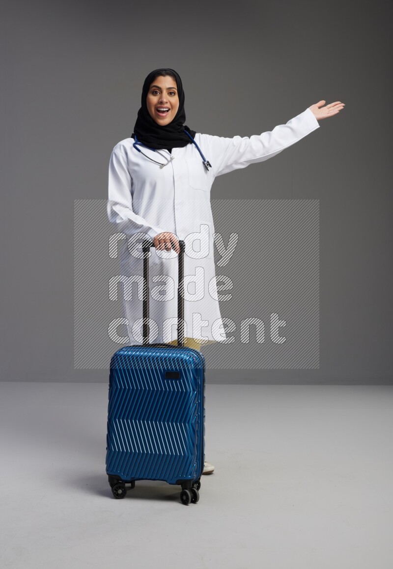Saudi woman wearing lab coat with stethoscope standing holding Travel bag on Gray background
