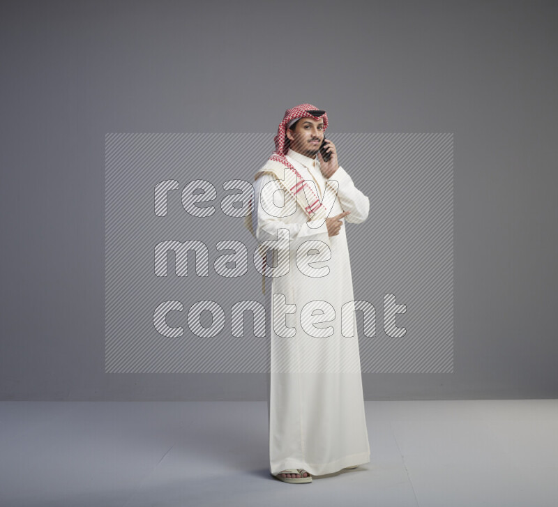 A Saudi man standing wearing thob and red shomag talking on phone on gray background