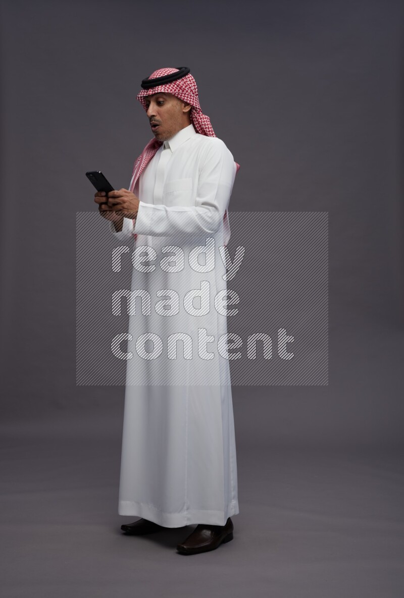 Saudi man wearing thob and shomag standing texting on phone on gray background
