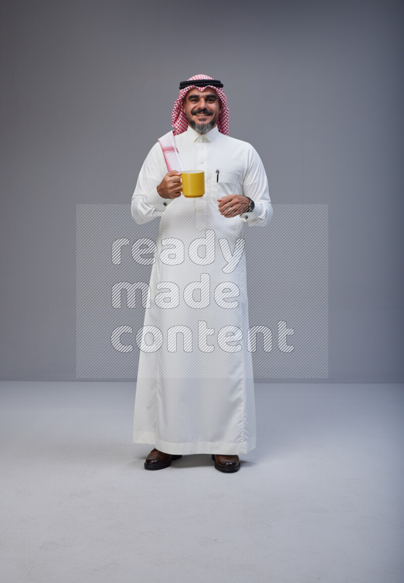 Saudi man Wearing Thob and red Shomag standing holding mug on Gray background