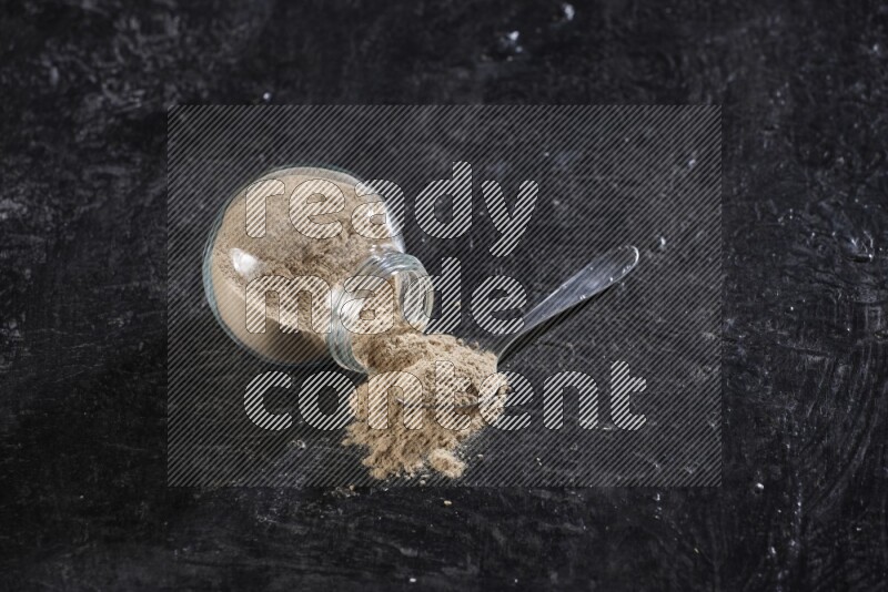 A glass spice jar full of garlic powder flipped and the powder came out with a filled metal spoon beside it on a textured black flooring