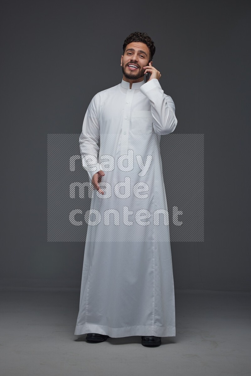 A Saudi man wearing Thobe and talking in the phone while standing and making different hand gestures eye level on a gray background