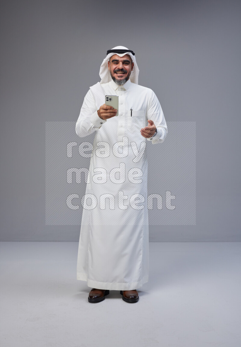 Saudi man Wearing Thob and white Shomag standing texting on phone on Gray background
