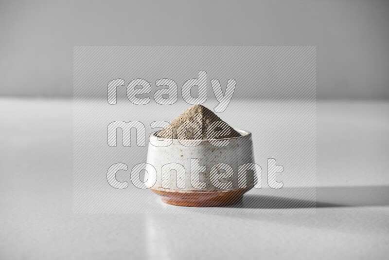 A white ceramic bowl full of black pepper powder and black pepper beads spread on white flooring