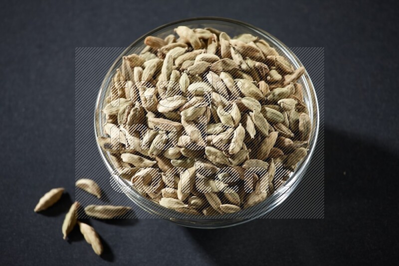 A glass bowl full of cardamom and more seeds spreaded beneath the bowl on black flooring