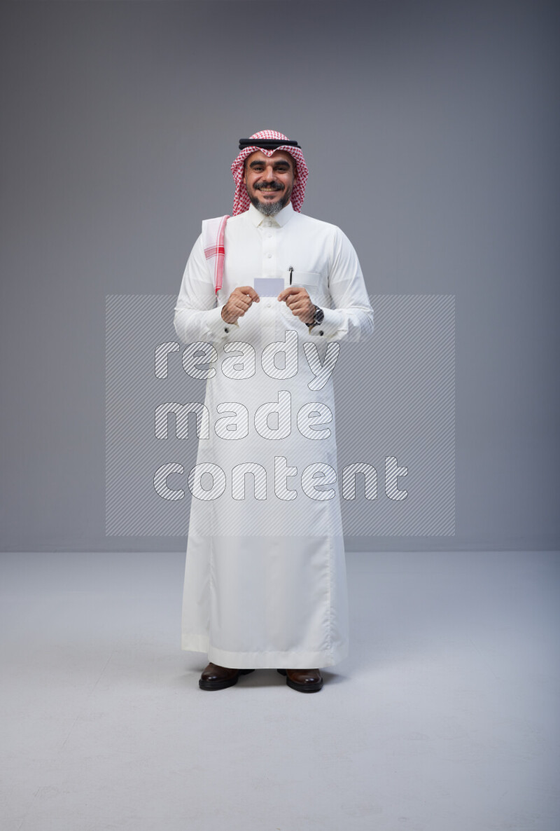Saudi man Wearing Thob and red Shomag standing holding ATM card on Gray background