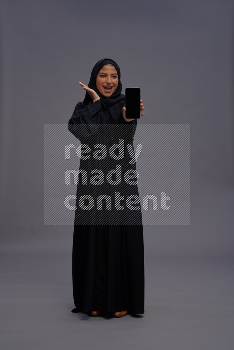 Saudi woman wearing Abaya standing showing phone to camera on gray background