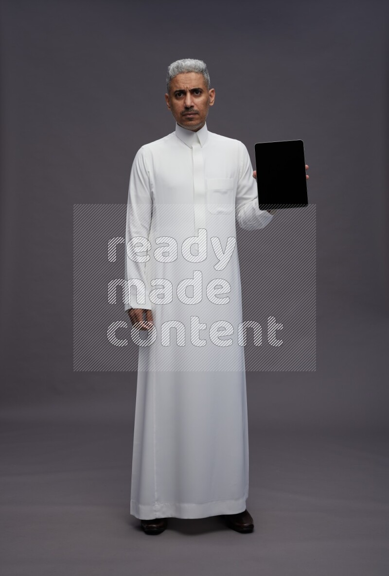 Saudi man wearing thob standing showing tablet to camera on gray background