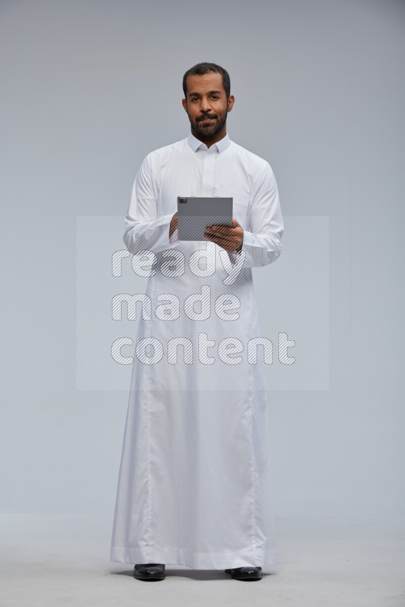 Saudi man wearing Thob standing working on tablet on Gray background
