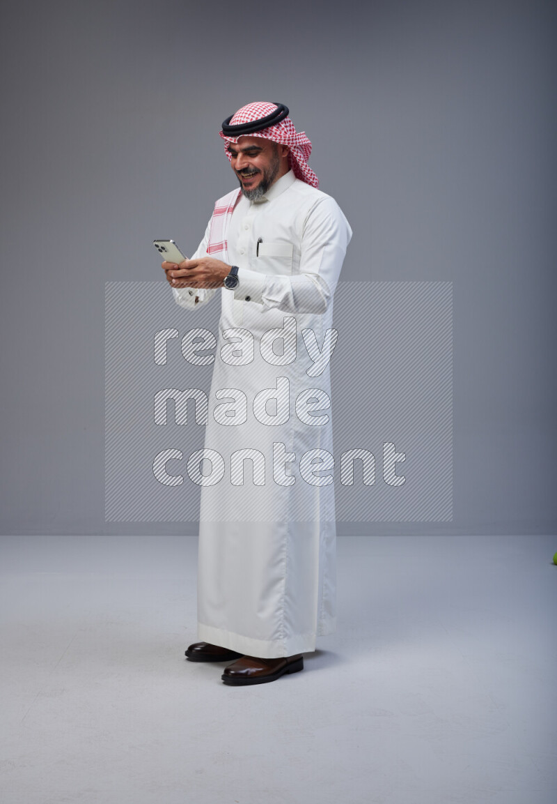 Saudi man Wearing Thob and red Shomag standing texting on phone on Gray background
