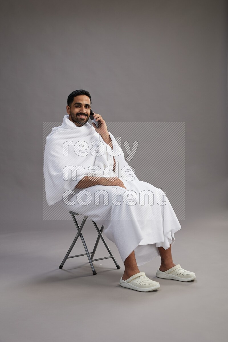 A man wearing Ehram sitting on chair talking on the phone on gray background