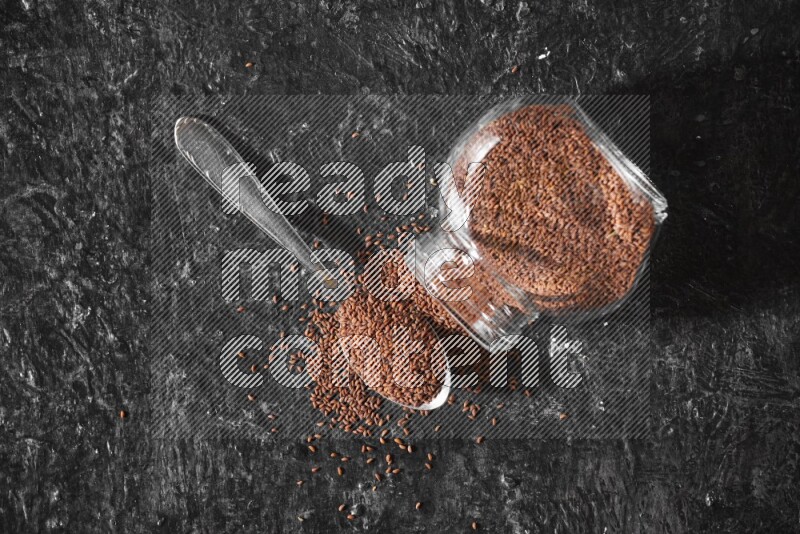A glass spice jar full of garden cress seeds flipped and seeds spread out with a metal spoon full of the seeds on a textured black flooring