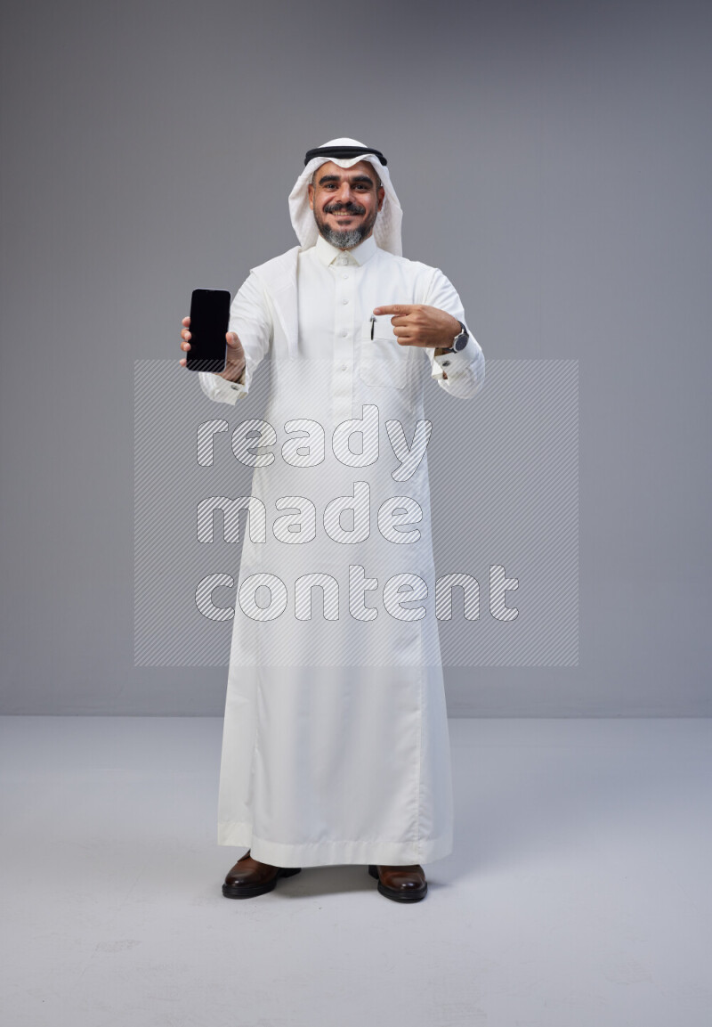 Saudi man Wearing Thob and white Shomag standing showing phone to camera on Gray background