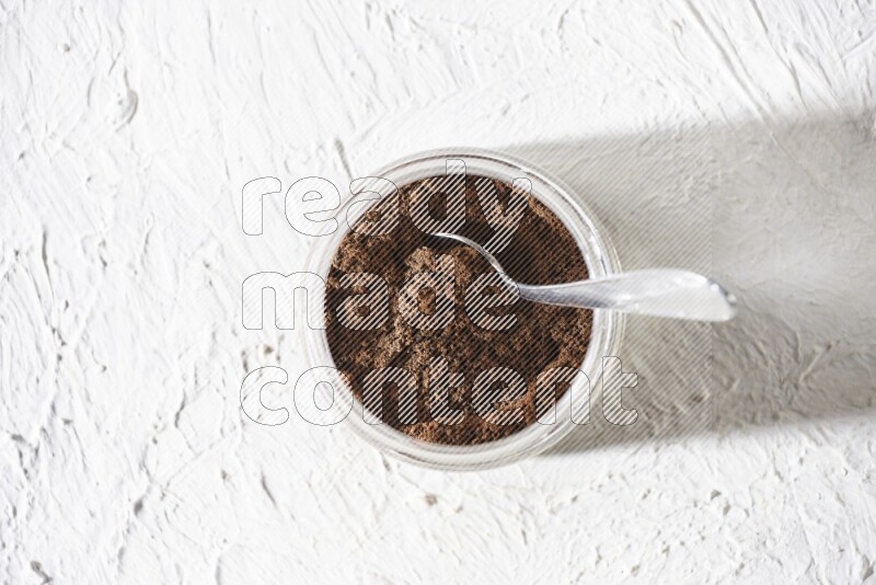 A glass jar full of cloves powder with a metal spoon on a textured white flooring