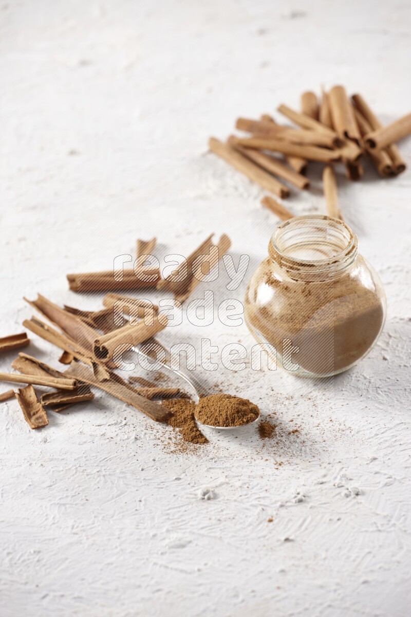 Herbal glass jar full cinnamon powder and a metal spoon surrounded by cinnamon sticks on a white background