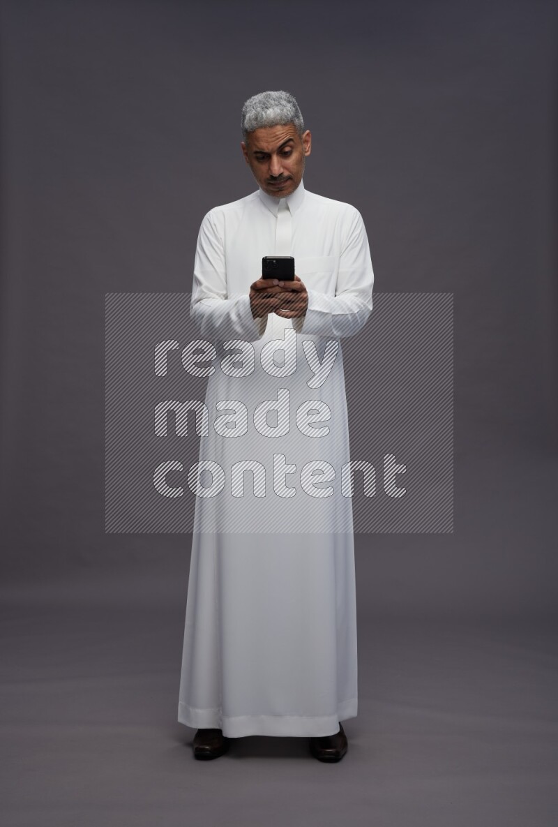 Saudi man wearing thob standing texting on phone on gray background