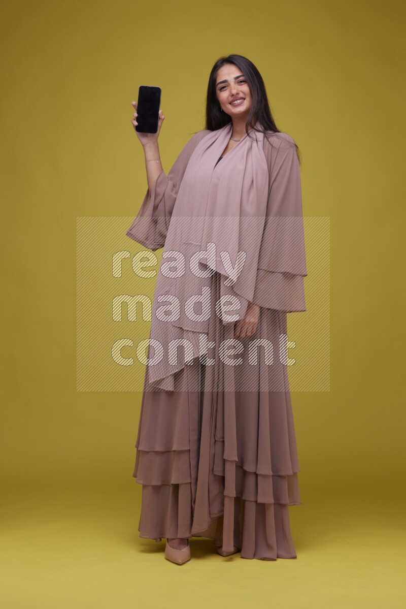 A woman Showing SmartPhone Screen on a Yellow Background wearing Brown Abaya