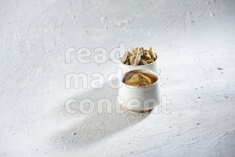 وعاءان فخاريان بيج ممتلئان ببودرة الكركم وأصابع الكركم المجففة على خلفية بيضاء