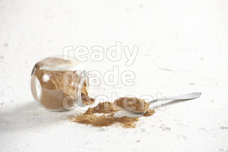 Flipped herbs glass jar full of cinnamon powder with a metal spoon full of powder on a textured white background