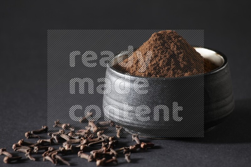 A black pottery bowl full of cloves powder and cloves whole grains on a black flooring
