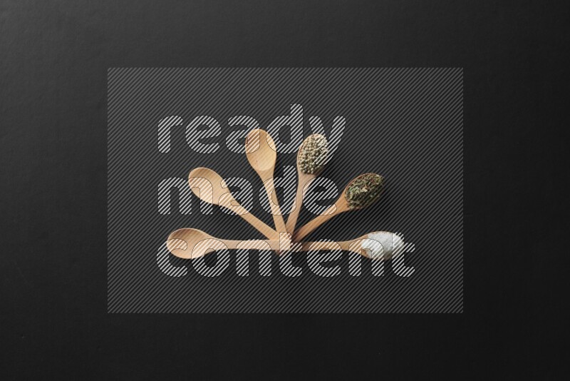 wooden spoons filled with white peppers, cloves, cardamom, salt, black peppers and basil on black flooring