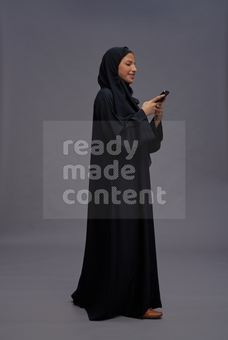 Saudi woman wearing Abaya standing texting on phone on gray background