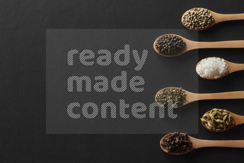 wooden spoons filled with white peppers, cloves, cardamom, salt, black peppers and basil on black flooring