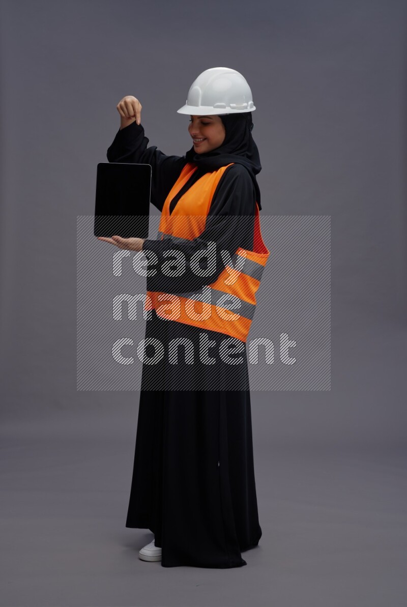 Saudi woman wearing Abaya with engineer vest standing showing tablet to camera on gray background