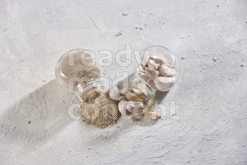 2 glass spice jars, one full of garlic powder and the other full of garlic cloves both flipped on a textured white flooring