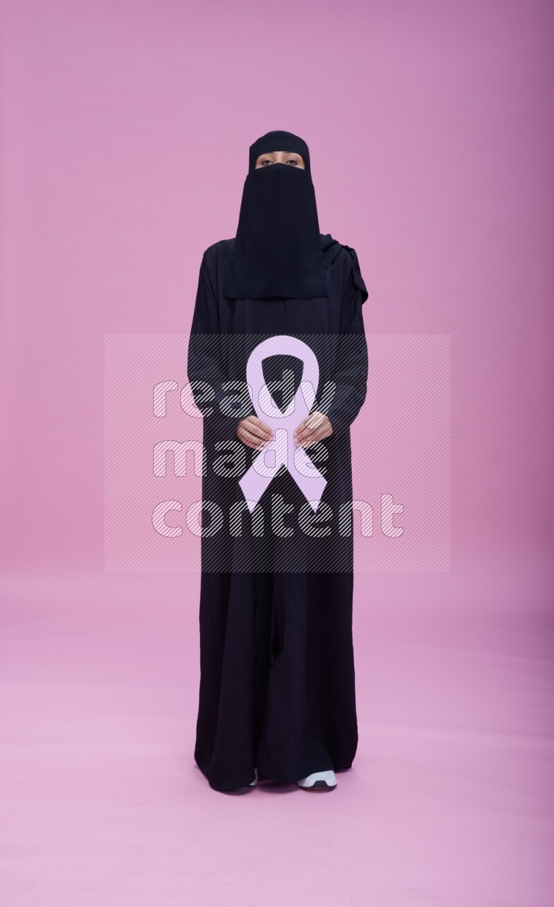 Saudi woman wearing abaya and niqab standing holding awareness ribbon on pink background