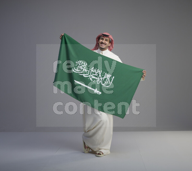 A saudi man standing wearing thob and red shomag holding big saudi flag on gray background