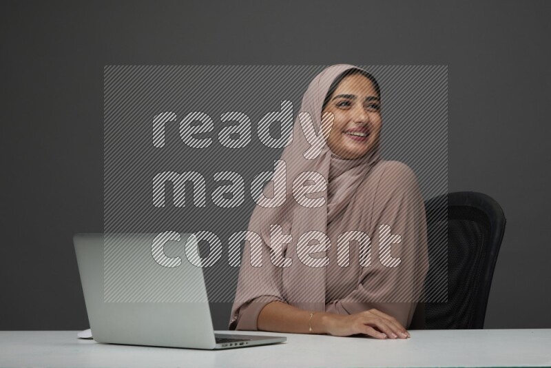 A Saudi woman Setting on her desk on a Gray Background wearing Brown Abaya with Hijab