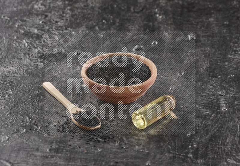 A wooden bowl and spoon full of black seeds and a bottle of black seeds oil on a textured black flooring