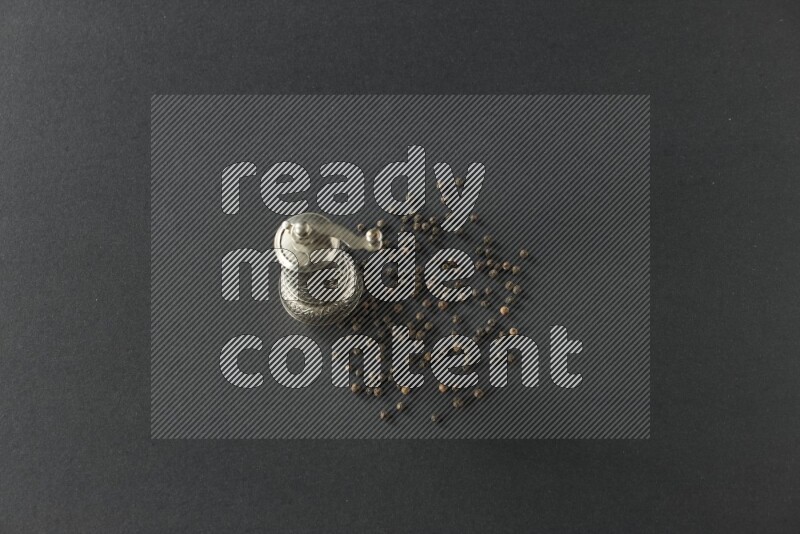 مطحنة فلفل تركية معدنية وحبوب فلفل أسود متناثرة على أرضية سوداء